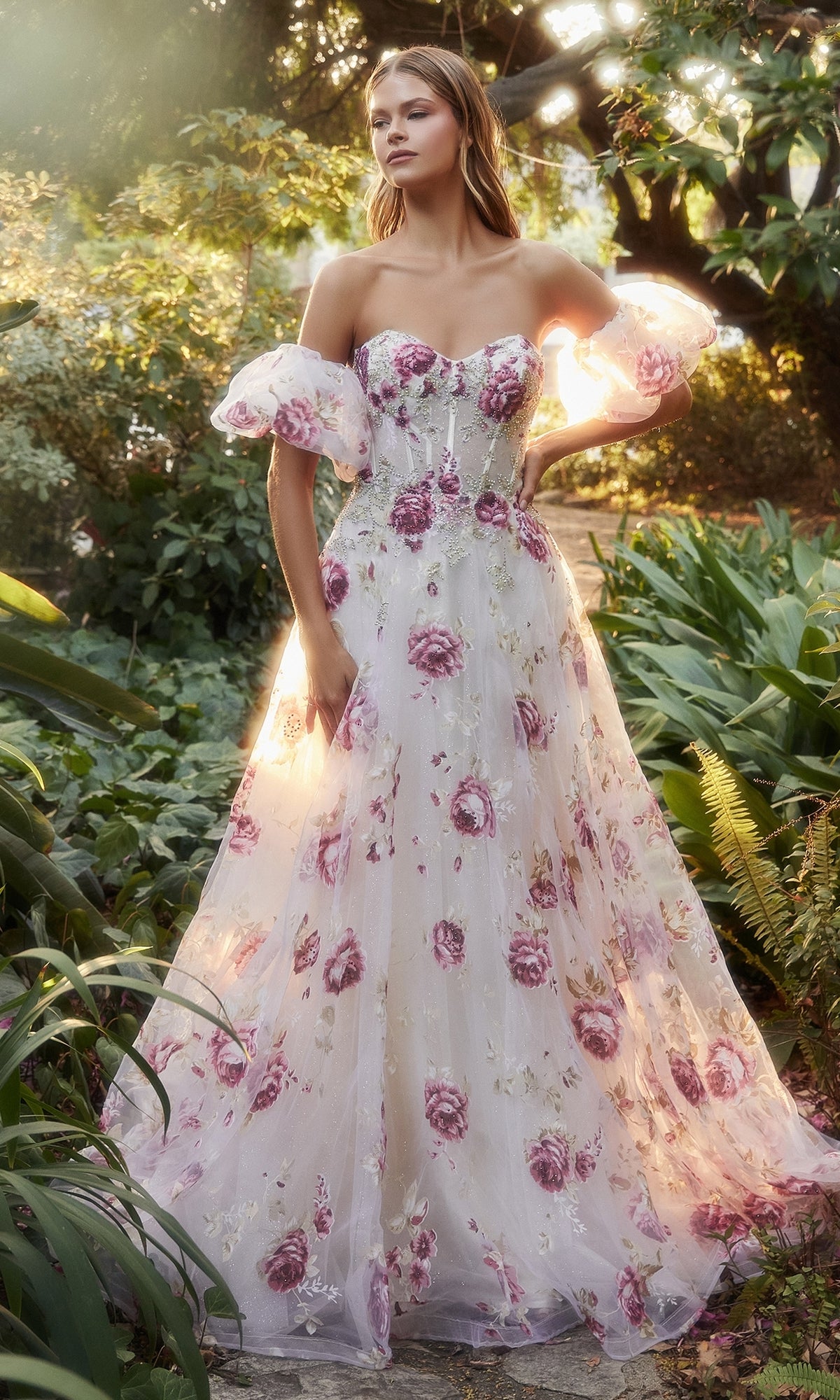 A woman stands outdoors in the Puff-Sleeve Strapless Floral-Print Ball Gown A1133, featuring a white strapless bodice with delicate pink patterns and sheer puff sleeves, surrounded by lush green plants and sunlight.